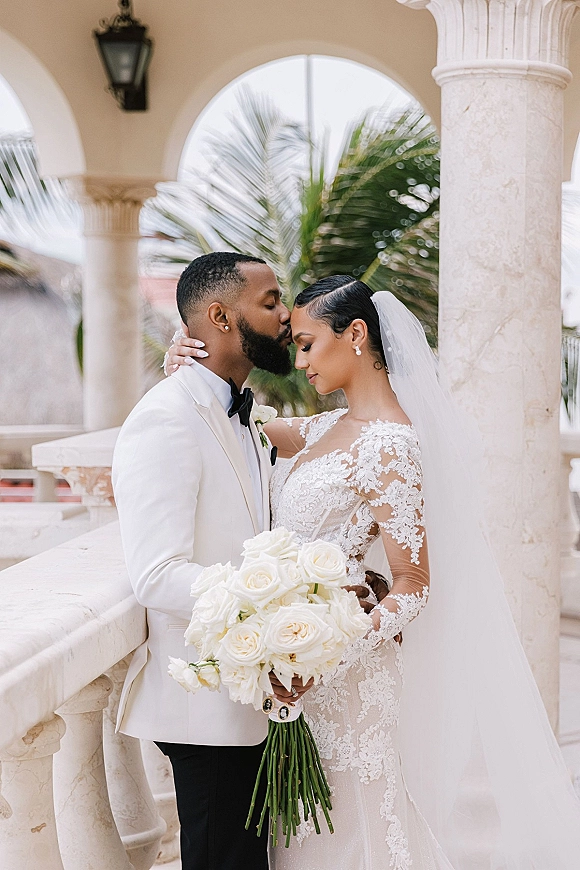 Wedding couple portrait of bride and groom forehead kiss, bride holding white rose bouquet under stone columns on an arched veranda