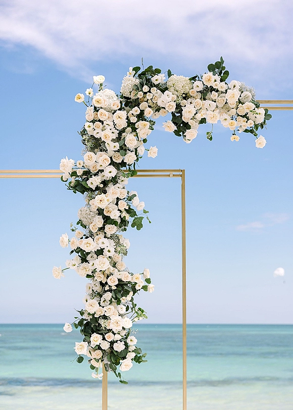 Wedding arch flowers on a gold square wedding arch with white roses and hydrangeas, greenery accents, set against ocean and blue sky backdrop