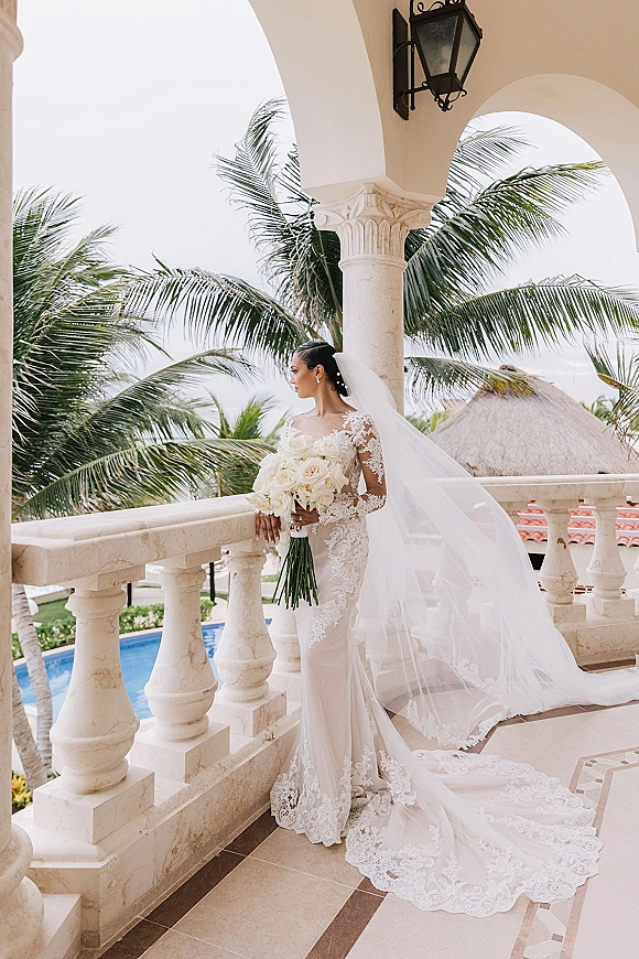 Bridal portrait of bride holding bouquet in a lace wedding dress with long veil, leaning on stone balcony at a tropical resort terrace with palms