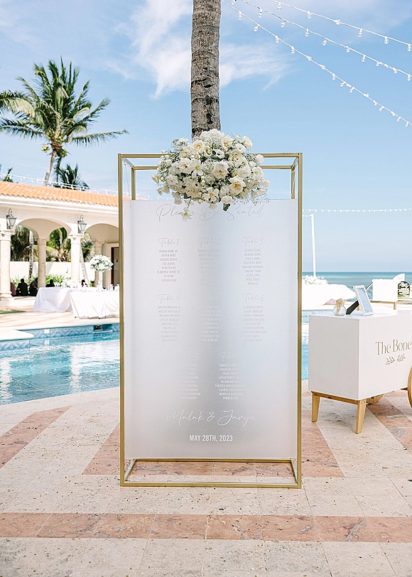 Wedding seating chart in a gold frame, seating chart sign topped with white florals and string lights on a poolside patio by the ocean