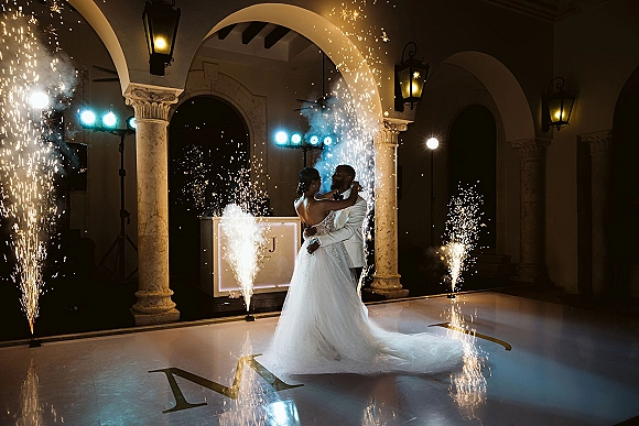 First dance with sparkler first dance fountains as bride in gown with long train and groom in tuxedo sway on monogram floor in columned hall