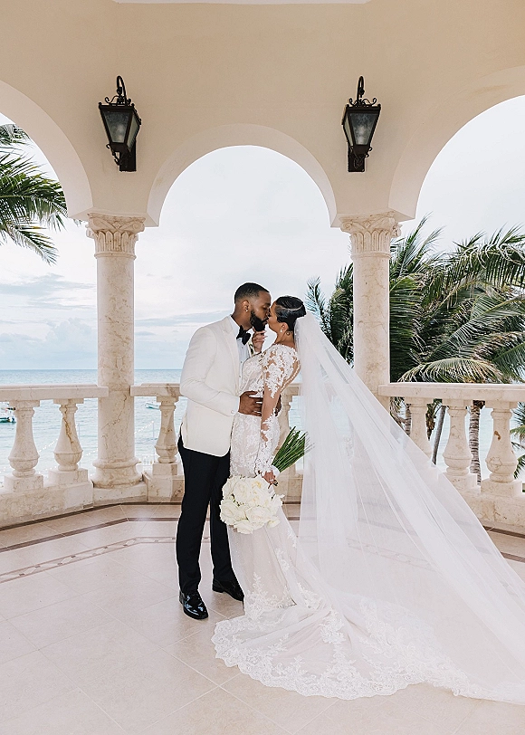 Wedding kiss portrait of bride and groom kissing under a stone archway, her cathedral veil flowing, holding a white bouquet with ocean beyond