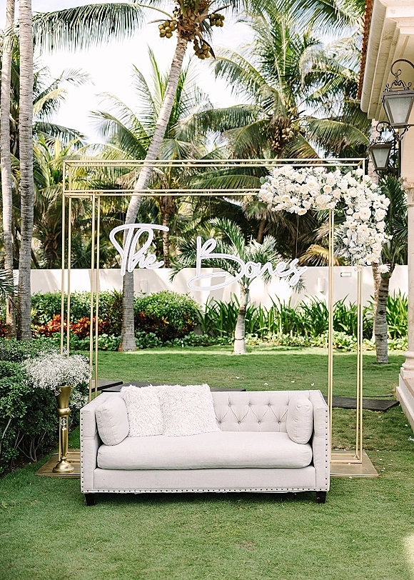 Wedding photo backdrop with a gold metal frame, white florals, and a custom neon sign beside a white sofa in a palm-lined courtyard.