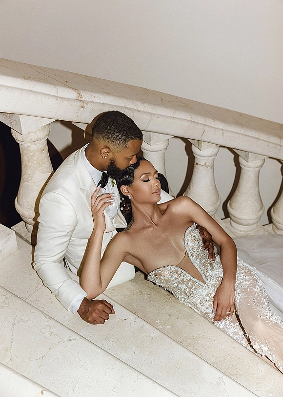 Couple portrait of groom kissing bride’s forehead as she reclines on a marble staircase, wearing a strapless lace wedding dress with pearl hair pins