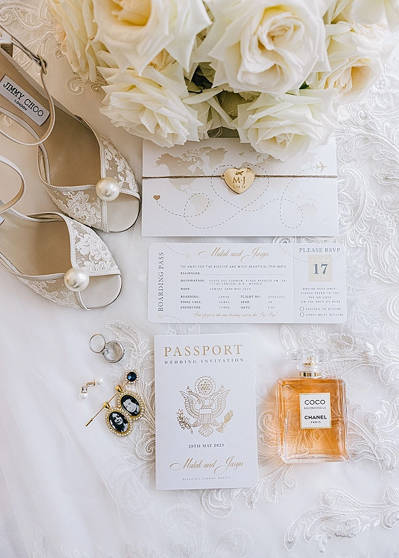 Invitation suite with destination wedding invitations, passport and boarding pass cards, rings, pearl shoes, and rose bouquet on lace fabric