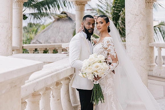 Couple portrait of bride in lace gown and cathedral veil holding a white bouquet, groom in tuxedo, on a stone balcony with palms.