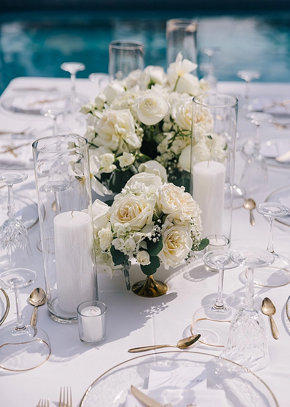 Wedding tablescape with a white rose centerpiece, greenery, gold flatware, and taper candles on a waterfront outdoor reception table in daylight