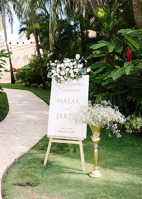 Wedding welcome sign on an easel with white roses and eucalyptus in a gold pedestal vase along a tropical garden stone walkway