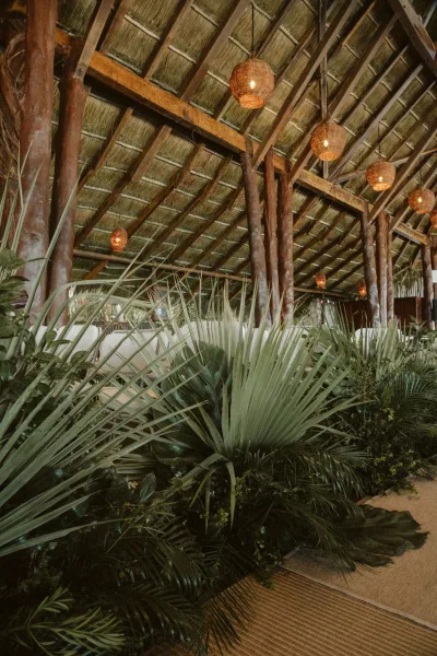 Reception lounge decor with tropical wedding greenery, woven pendant lights, and palm leaves under a thatched open-air pavilion ceiling