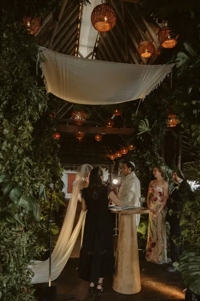 Wedding ceremony with couple exchanging vows under a fabric canopy, bride in black gown and long veil in a wood-beam pavilion with greenery backdrop