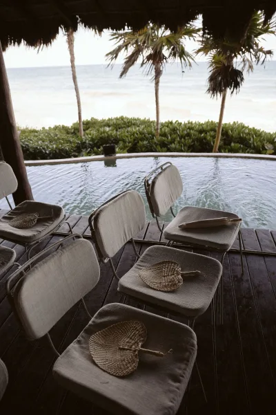 Ceremony seating with folding chairs and woven hand fans on cushions along a wooden deck, overlooking an ocean view with palms by the pool
