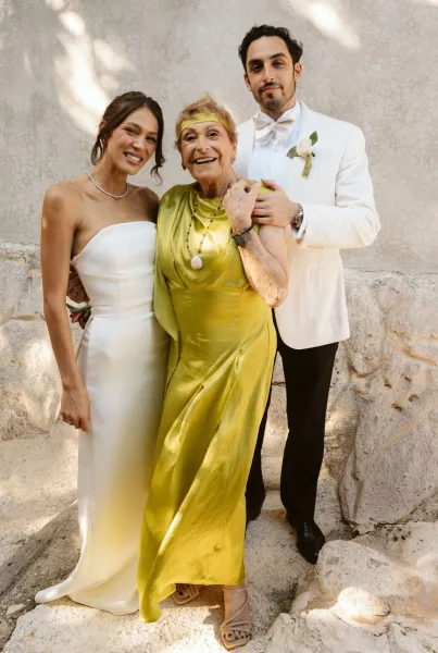 Wedding family portrait of bride in strapless gown, groom in white tuxedo, and grandmother in green satin dress by a sunlit stone wall