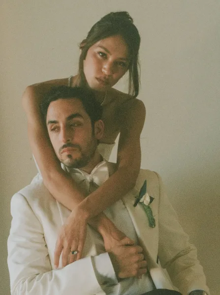 Couple portrait with bride hugging groom in a white tuxedo jacket and black bow tie, her strapless gown and jewelry against a plain wall