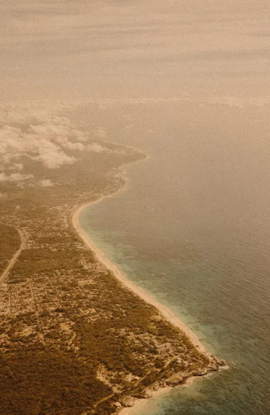 Aerial coastline view of ocean shoreline and sandy beach curving past a coastal town, with clouds and hazy sky along the horizon