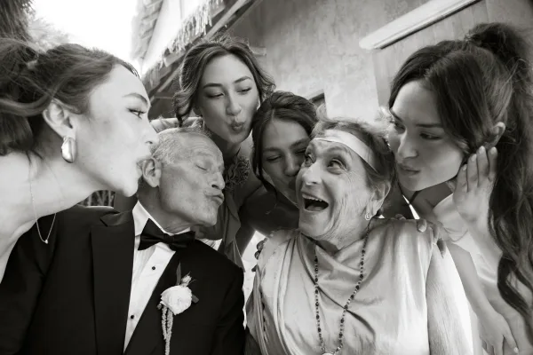 Wedding family photo of bride and groom with grandparents as the group kisses cheeks, in daylight by an outdoor building wall, tuxedo accents