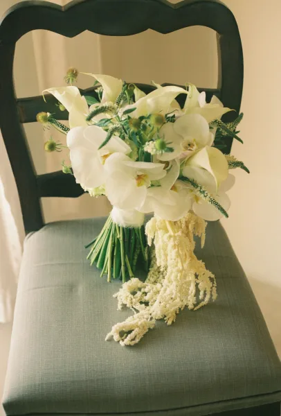 Bridal bouquet with white orchid bouquet blooms, calla lilies, and hanging amaranthus draped over a dark wooden chair by sheer curtains