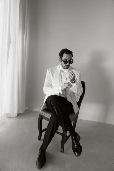 Groom portrait in a white tuxedo adjusting cufflinks, wearing sunglasses and a bow tie, seated by sheer-curtained window light