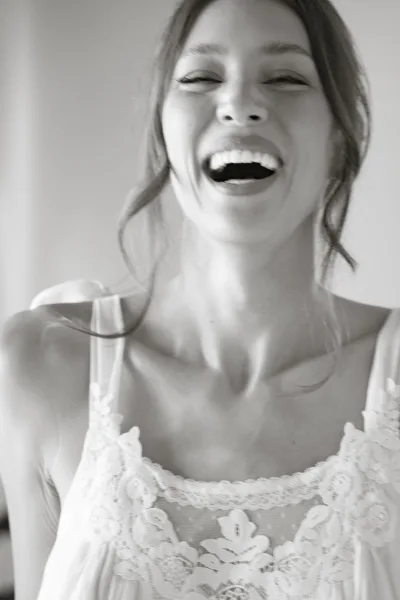 Bridal portrait of a laughing bride in a lace wedding dress with spaghetti straps, black and white against a plain wall backdrop