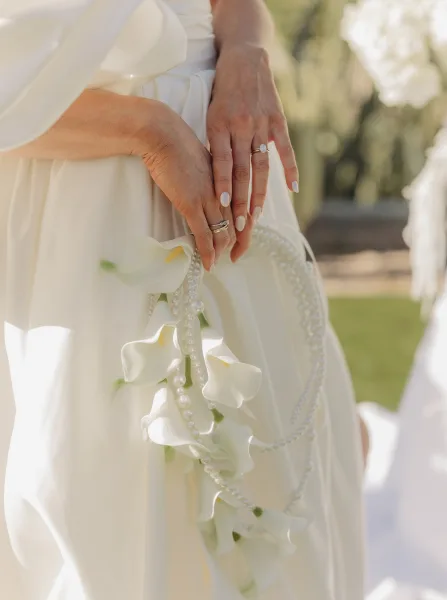 Bridal ring close-up showing engagement ring close up and wedding band stack on manicured hand with pearl strands, sunlit greenery bokeh
