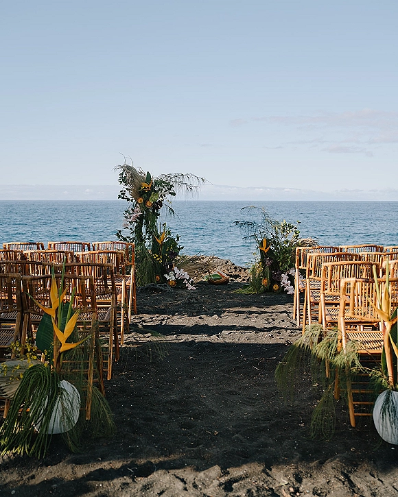Beach ceremony setup with oceanfront wedding ceremony aisle, bamboo chairs and a tropical floral arch on a black sand beach by rocks