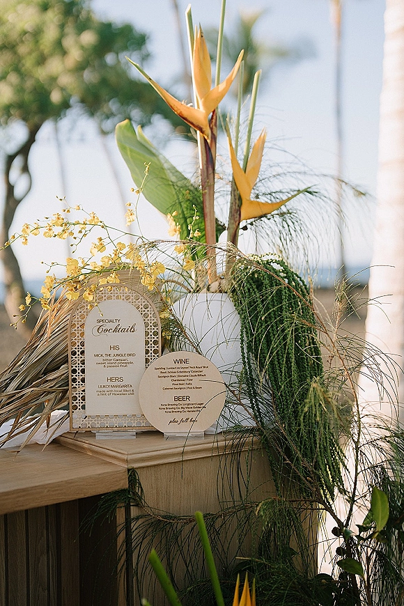 Wedding bar signage with a signature cocktail sign and menu cards on acrylic stands, styled with bird of paradise on a coastal patio backdrop