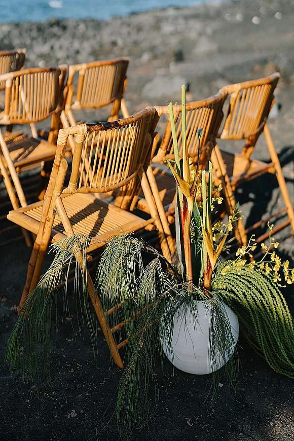 Ceremony seating with bamboo wedding chairs arranged by a rocky shoreline, accented with tropical foliage and a white lantern by the aisle