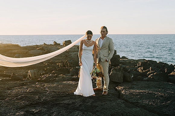 Couple portrait of a beach wedding couple on a rocky shoreline, bride in slip dress with long veil and bouquet beside groom in tan suit