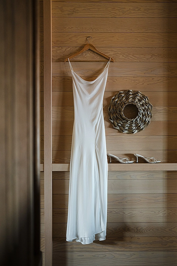 Wedding dress hanging on a wooden hanger with strappy heels below against a wood panel wall, minimalist bridal details scene