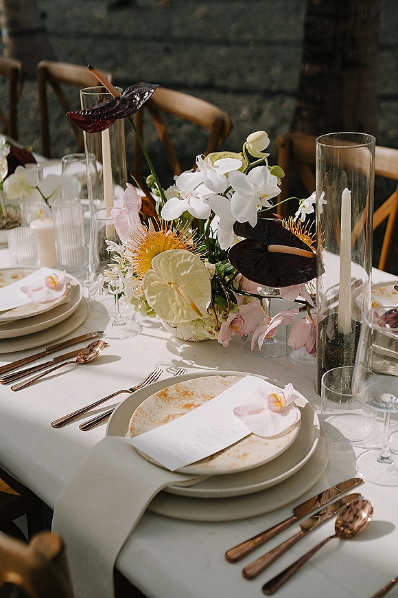 Reception tablescape wedding place setting with anthurium and orchid centerpiece, taper candles, rose gold flatware, against a stone wall backdrop