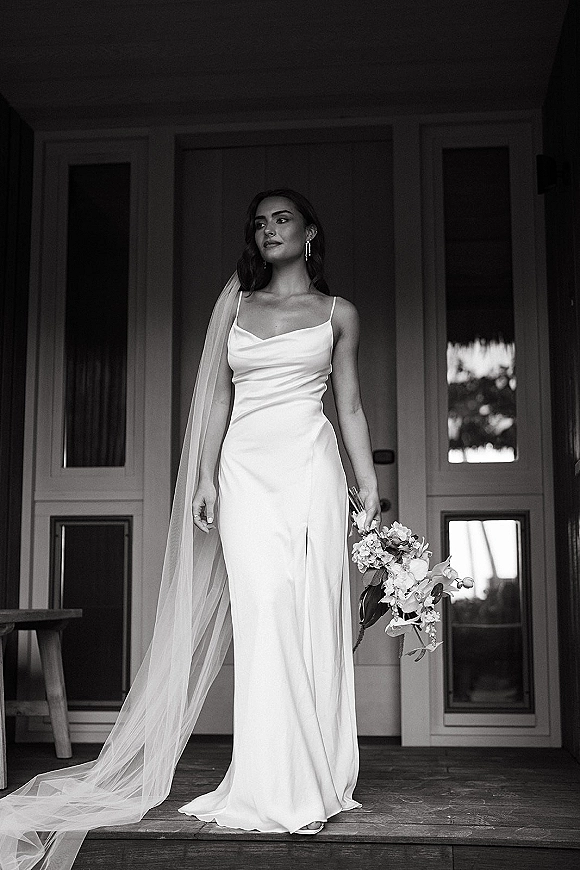 Bridal portrait in black and white of a bride holding a white orchid bouquet, wearing a long veil and drop earrings on a porch with paneled doors
