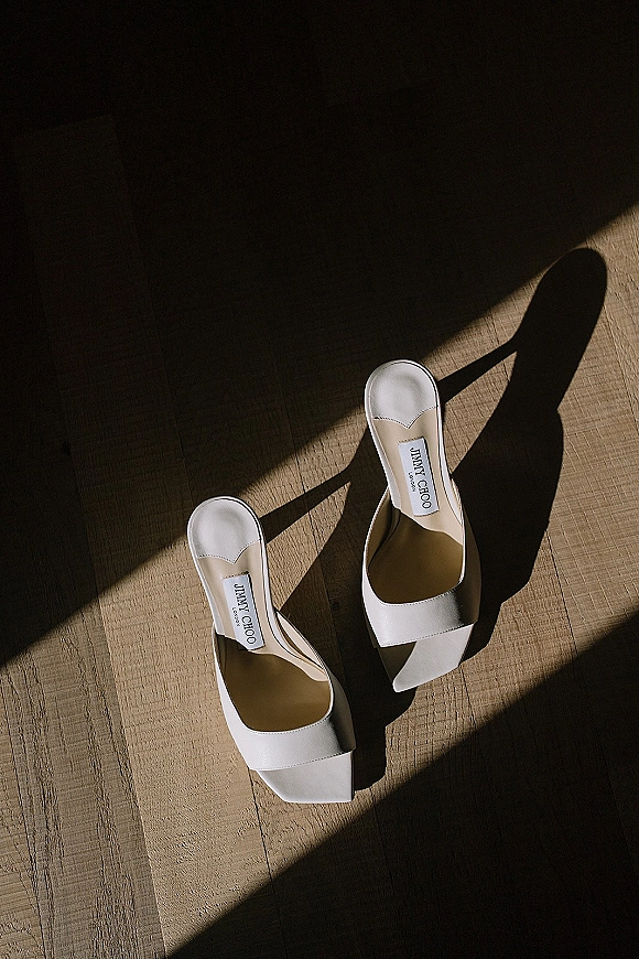 Bridal shoes, white wedding heels with ankle straps and open toes, resting on a wood floor with soft natural light shadows