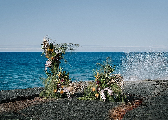 Ceremony floral arrangement with beach wedding florals of orchids and palm fronds on seaside rocks against ocean and blue sky