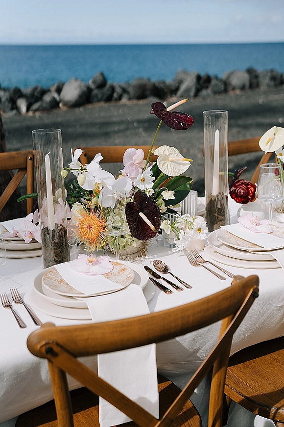 Reception tablescape with a coastal wedding tablescape of orchids, anthurium, and pillar candles in glass vases beside ocean and rocky shore
