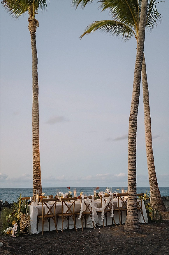 Reception tablescape with a beach reception table featuring white linens, taper candles, floral garland, and crossback chairs by the oceanfront sky