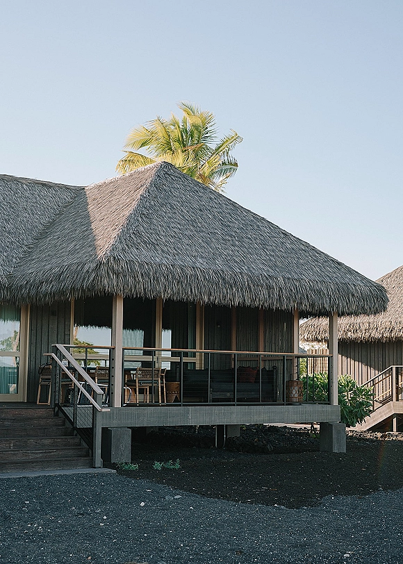 Wedding venue exterior with a thatched roof and wooden deck stairs, glass doors, and outdoor seating beside palm trees under a clear sky