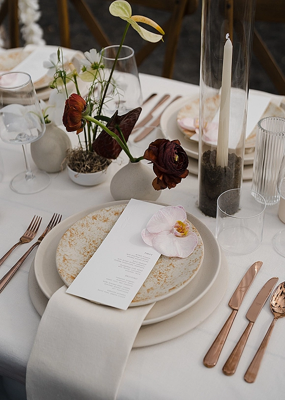 Reception tablescape with wedding place setting, ceramic plates topped with menu card and orchid, rose gold flatware, taper candle, bud vases on white linens