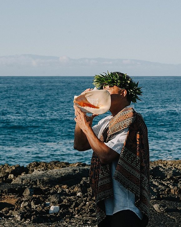 Wedding conch shell ceremony as officiant blows a conch, wearing a leaf crown and patterned shawl on a rocky ocean shoreline.