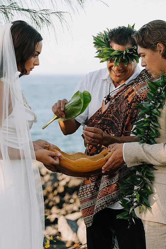 Ceremony moment during a wedding blessing ritual as officiant holds a wooden bowl with a green leaf, couple in leis by rocky ocean shore