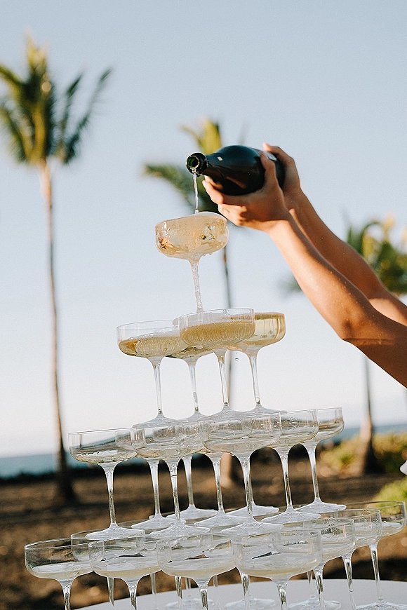 Champagne tower of stacked coupe glasses with champagne bottle nearby, set outdoors with palm trees and a distant ocean backdrop