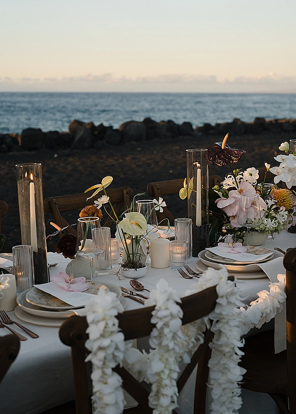 Reception tablescape with candlelit chargers, menus, orchids and anthurium centerpiece, white lei chairs, set beside an oceanfront rocky shore