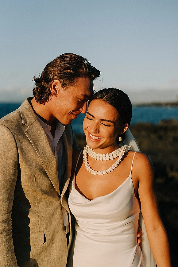 Couple portrait of newlyweds in a beach wedding couple moment, bride in veil and shell lei laughing as groom leans close by ocean sky