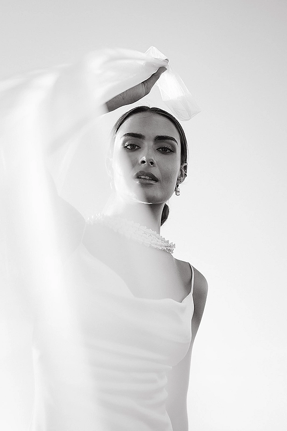 Bridal portrait in black and white with veil over face, satin slip wedding dress and pearl necklace against a minimalist studio backdrop