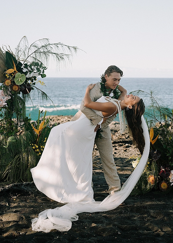 Wedding couple portrait of groom dipping bride in a white dress and long veil on a black sand beach with waves and tropical florals