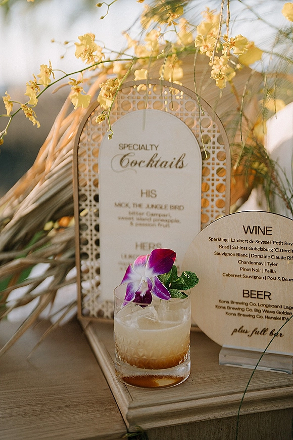 Wedding bar signage with specialty cocktails sign on wooden stands beside a rocks glass cocktail with orchid and mint in soft outdoor light