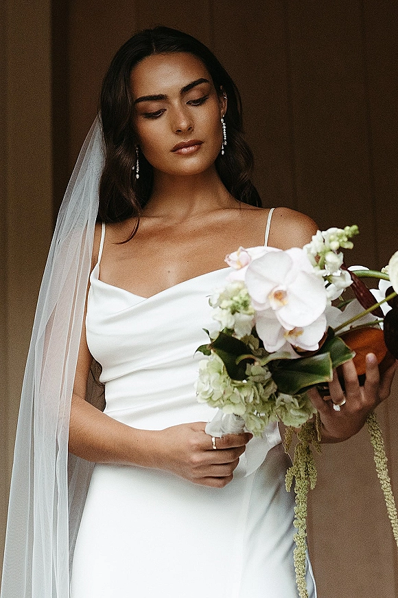 Bridal portrait of a bride holding bouquet with a long wedding veil, satin cowl-neck dress, and cascading white orchid bouquet on a neutral backdrop
