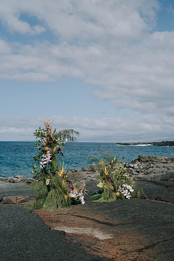 Ceremony floral arrangements with tropical greenery and orchids, styled asymmetrically on a rocky shoreline with ocean horizon under clouds