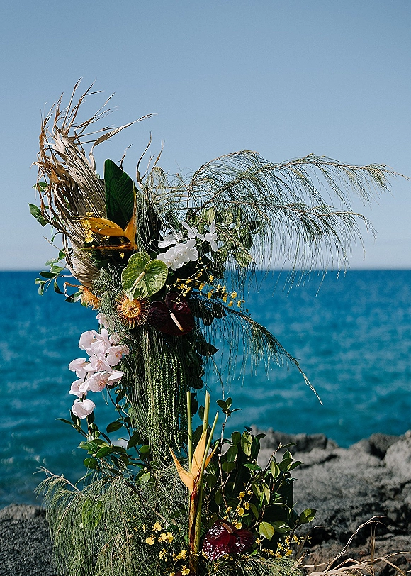 Wedding floral arrangement with tropical wedding flowers, orchids and palm fronds on a pedestal by an ocean rocky shoreline under blue sky