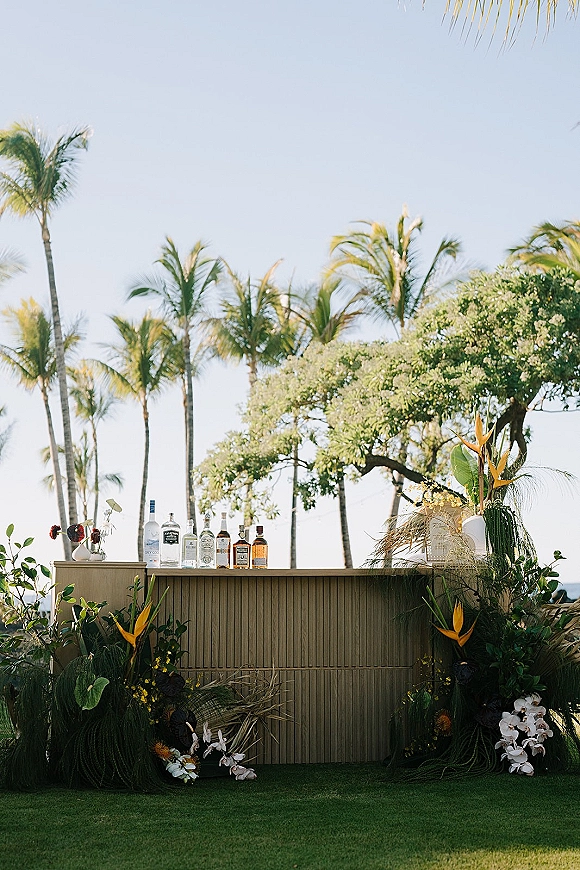 Wedding bar setup with liquor bottles and bird of paradise floral accents on a bar counter, set on a lawn with palm trees and ocean view