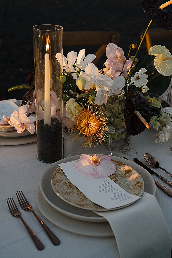 Reception tablescape with wedding place setting, menu card on ceramic plate, orchid centerpiece and hurricane candle on white linen outdoors