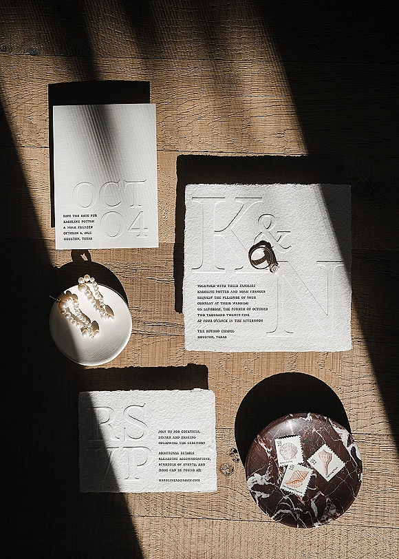 Invitation suite with letterpress wedding invitations, RSVP and save the date, wedding rings and pearl earrings on a wood tabletop in window light